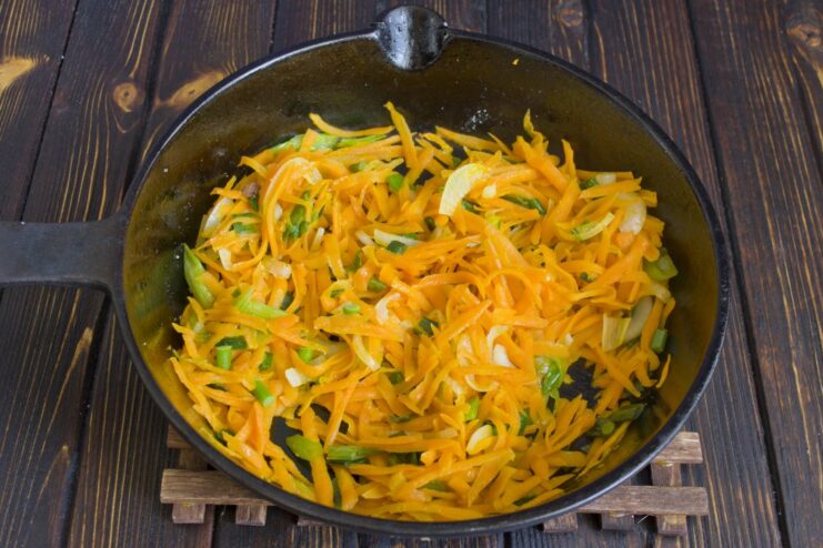 Adding grated carrots to the sautéed onions in the pan. Grated carrots being fried together with chopped onions in a pan.