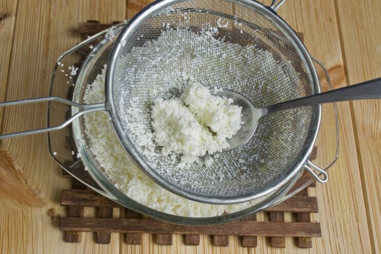 Sifting farmer's cheese through a sieve to achieve a smooth, lump-free texture. Farmer's cheese being pressed through a fine-mesh sieve into a white bowl.