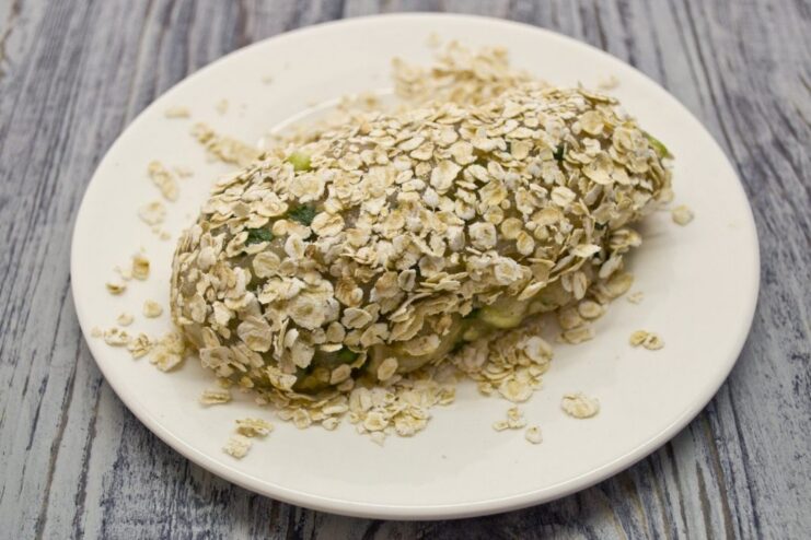Coating the Stuffed Cutlets in Rolled Oats A formed raw chicken cutlet stuffed with spinach and cheese being coated in rolled oats.