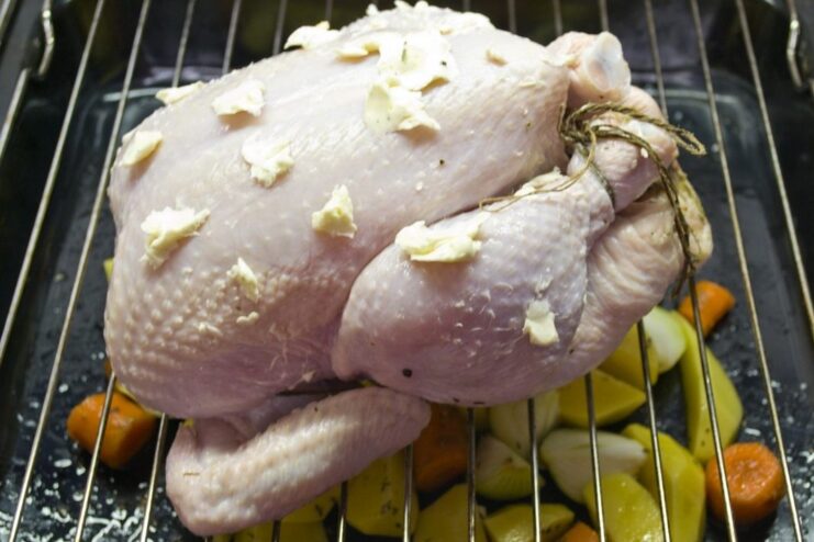 Roasting chicken on a rack allows juices to flavor the vegetables below. A whole chicken on a roasting rack placed over a pan of chopped vegetables, ready for the oven.