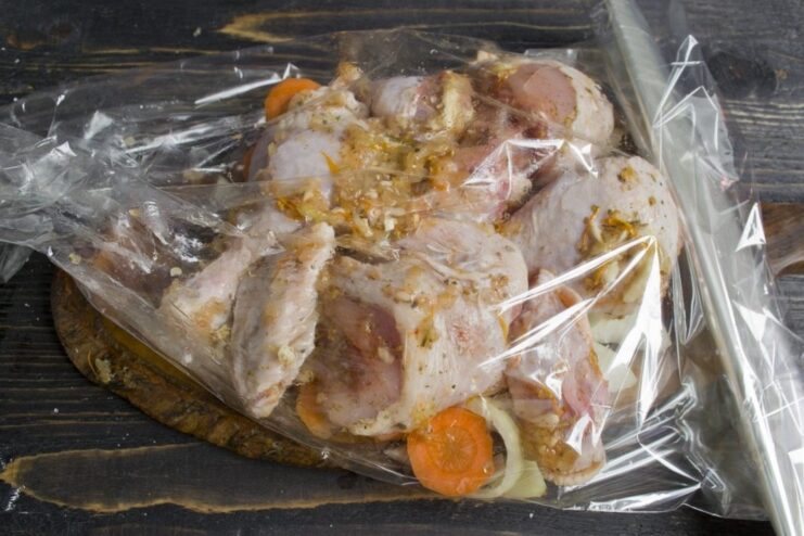 Assembling the chicken and vegetables inside the oven bag before baking. Marinated chicken being placed on a layer of onions and carrots inside a clear oven bag.