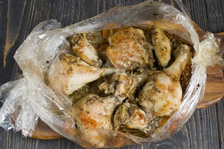 Chicken and vegetables roasting inside an inflated oven bag in the oven. An oven bag puffed up with steam, with chicken and vegetables visible inside, baking on a tray.