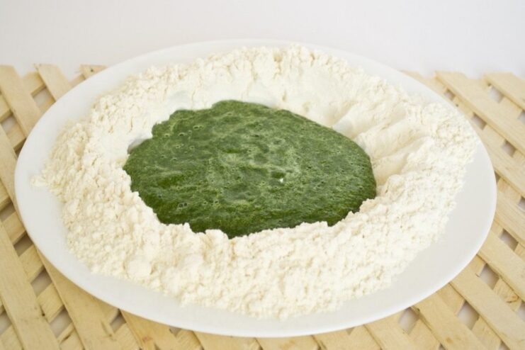 Step 4: Combining the spinach puree with flour to form the dough. Green spinach puree being mixed into a well of flour on a wooden board.