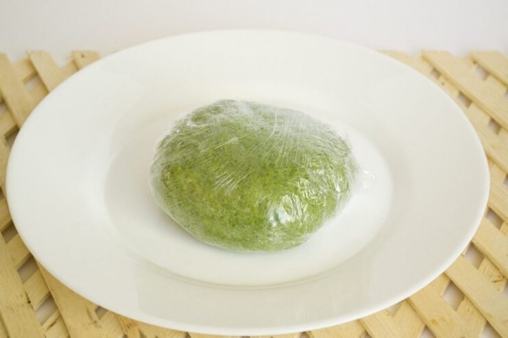 Step 5: Letting the dough rest to develop gluten and make it easier to roll. A ball of green spinach pasta dough wrapped in plastic wrap.