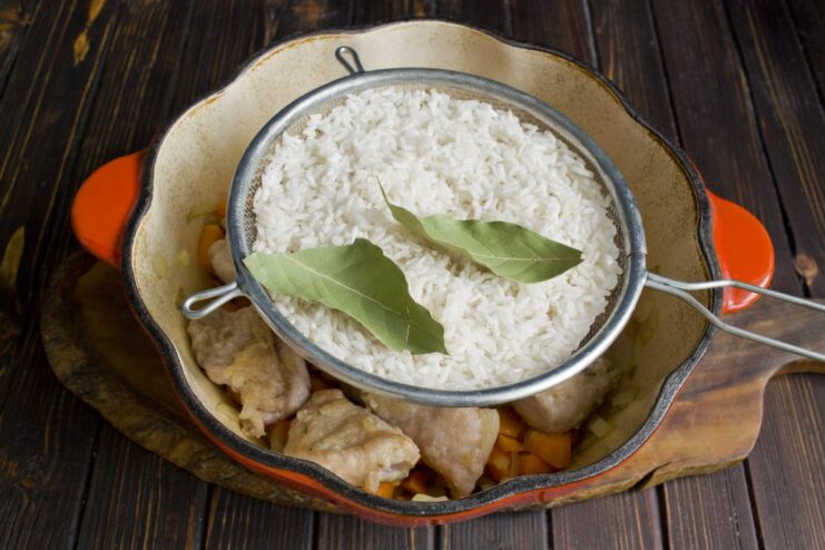 Adding a layer of rice and bay leaves over the pork. Uncooked white rice and bay leaves spread evenly over the pork and vegetables.
