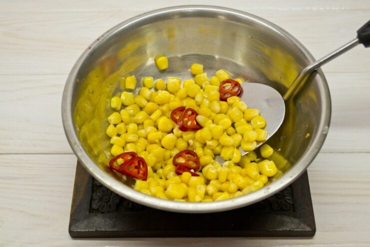 Adding sweet corn and spicy chili for extra flavor and color Canned corn and sliced chili pepper being added to the vegetable mix in a pan.