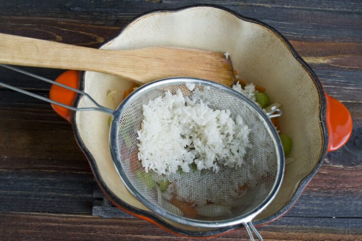 Step 3: Stirring in the rinsed jasmine rice to coat with flavors. Uncooked jasmine rice being added to a pan of sautéed vegetables.