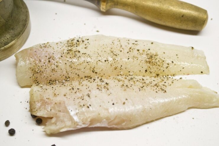 Seasoning the Fish Fillets Before Battering Raw fish fillets on a surface, being seasoned with salt and ground black pepper.