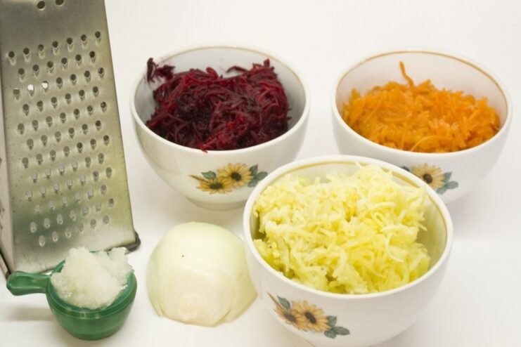 Using a fine grater to prepare the cooked beets, carrots, and potatoes for the salad layers. Cooked and peeled vegetables (beets, carrots, potatoes) being grated into separate bowls.