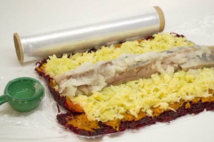 Assembling the Shuba Salad Roll: layering grated vegetables and herring on plastic wrap before rolling. Layers of grated beets, carrots, and potatoes with herring fillets arranged on top, ready to be rolled.