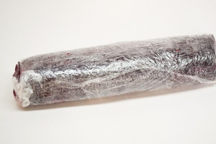 The final step before chilling: tightly rolling the layered salad using plastic wrap to form a perfect roulade. A person's hands tightly rolling the layered vegetable and herring salad into a log using plastic wrap.