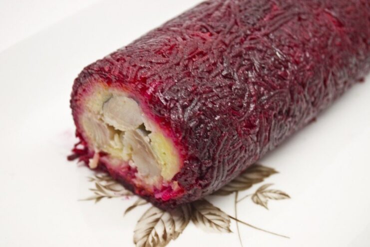 The finished Shuba Salad Roll, chilled and ready to be sliced and served as a stunning appetizer. The finished Shuba Salad Roll, a long log wrapped in plastic wrap, chilling on a plate.