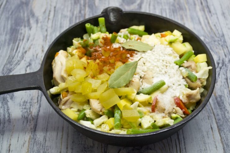 Adding Rice, Pickles, and Spices for the Signature Solyanka Flavor Arborio rice, chopped pickles, and spices being added to the vegetable and chicken mixture in the pan.