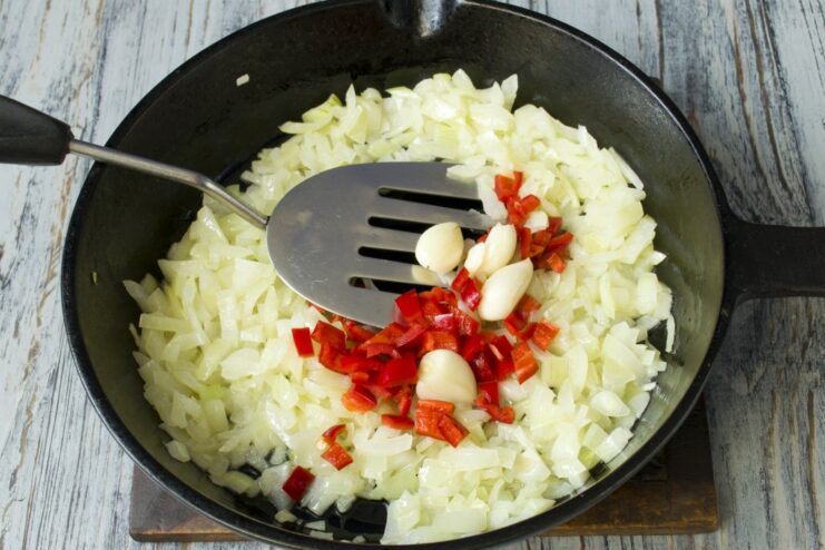 Adding aromatics: chili and garlic to the sautéed onion Sliced chili pepper and garlic cloves added to the sautéed onion in a pan.