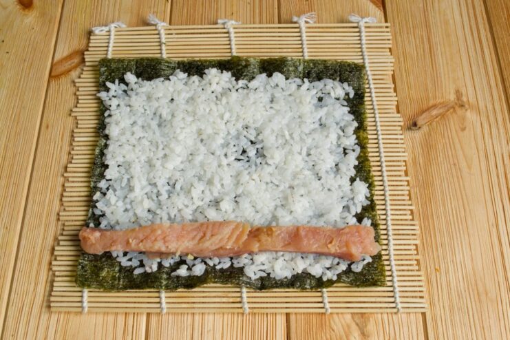 Adding the Filling: A Strip of Salted Red Fish for the Maki Roll A strip of bright orange salted salmon placed on the edge of the sushi rice.