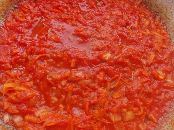 Tomato paste being added to sautéed onions and carrots in a pan.