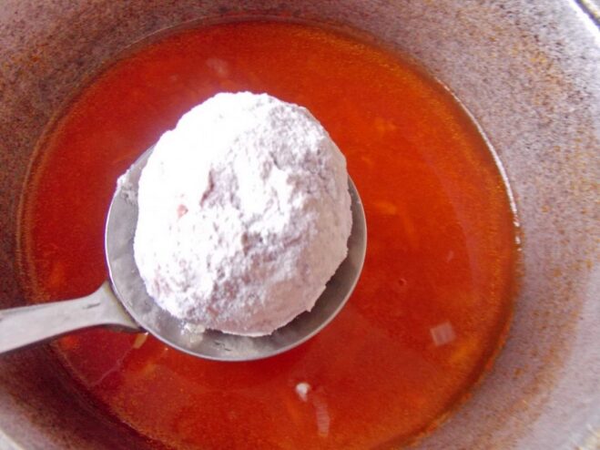 A raw meatball being dipped into a pot of boiling tomato sauce.
