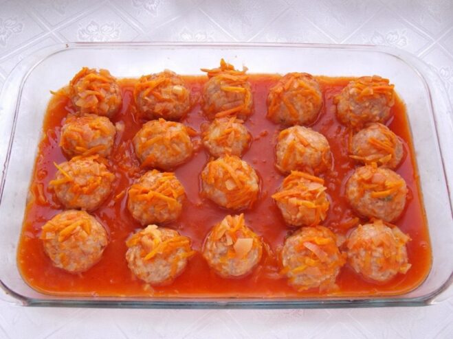 Tomato sauce being poured over raw meatballs in a baking dish.