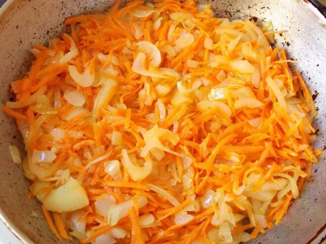 Adding grated carrots to the sautéed onions to build the flavor base. Grated carrots being added to the skillet with sautéed onions.