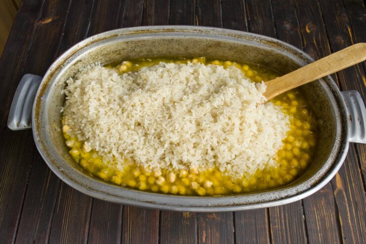 Spreading rinsed rice evenly over the other ingredients in the plov A layer of uncooked white rice being spread over the chicken and chickpeas.