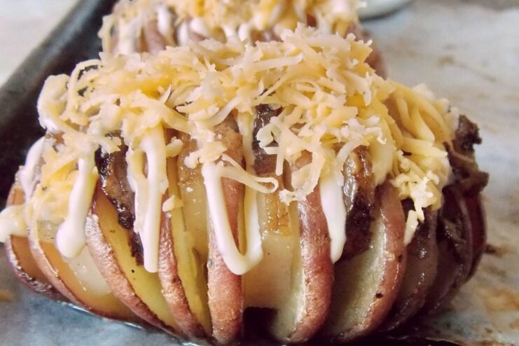 Topping the Baked Potatoes with Grated Cheese Baked accordion potatoes on a baking sheet being generously sprinkled with grated cheese before returning to the oven.