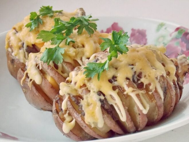 A close-up of golden-brown accordion potatoes with salo and melted cheese, garnished with fresh parsley on a baking sheet.