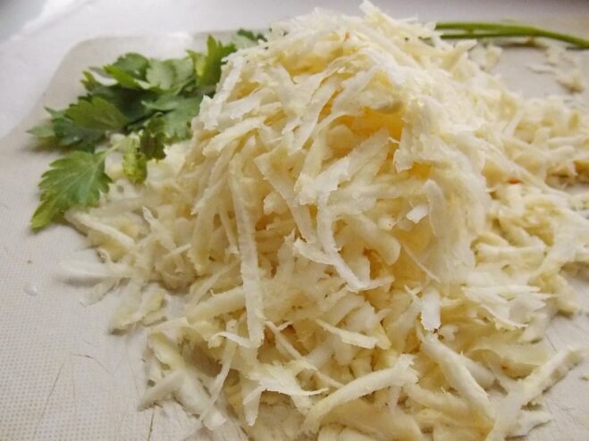 Grating celery root for the patties A hand grating a large celery root on a box grater into a bowl.