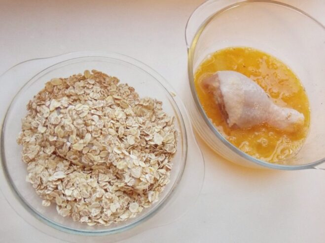 Step-by-step guide: coating a chicken drumstick in cereal flakes. A hand coating a raw chicken drumstick in a plate of cereal flakes after dipping it in egg.
