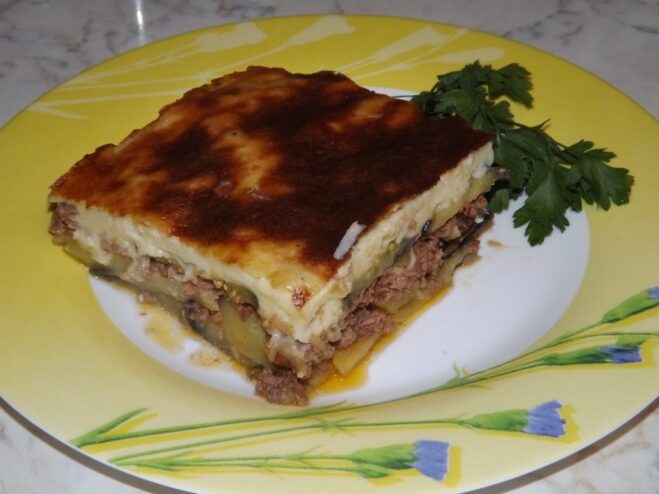 A square slice of Greek Moussaka on a white plate, showing layers of eggplant, meat, and a golden-brown béchamel topping.