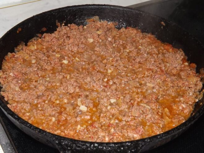Ground meat being added to the tomato and onion mixture in a pan.