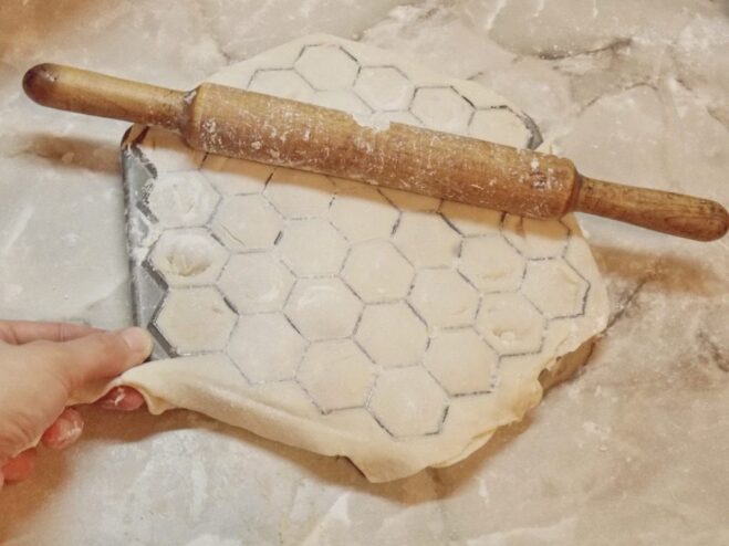 Sealing the Pelmeni with a Rolling Pin A second sheet of dough covers the filled pelmennitsa, with a rolling pin pressing down to seal the dumplings.