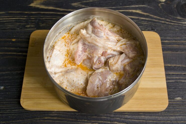 Chicken thighs soaking in a creamy smetana and spice marinade to tenderize the meat. Raw chicken thighs in a white bowl, coated with a creamy marinade of smetana and spices.