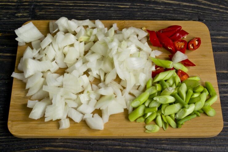 Preparing the aromatic base: chopped onion, garlic shoots, and chili pepper. Chopped white onion, green garlic shoots, and red chili pepper rings on a cutting board.