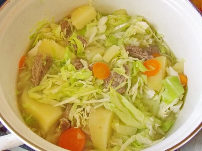 Adding shredded cabbage to the pot with potatoes and carrots. Shredded fresh cabbage being added to the pot of simmering vegetables for the stew.