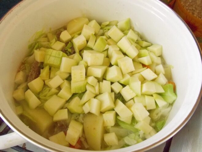 Adding diced young zucchini to the vegetable stew. Diced zucchini being added to the pot with other vegetables like cabbage, carrots, and potatoes.