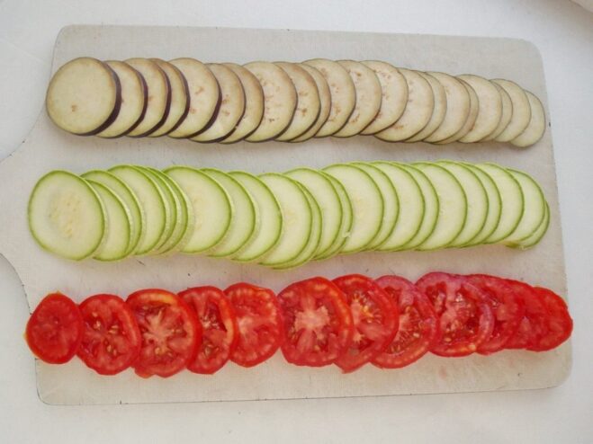 Preparing Vegetables for Ratatouille Tian Thinly sliced rounds of eggplant, zucchini, and tomato on a wooden cutting board.