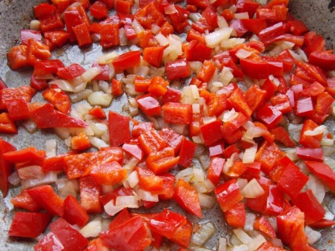 Adding Bell Peppers to the Sauce Base Diced red bell pepper being added to the sautéed onions in a pan.
