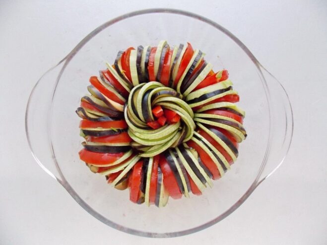 Arranging the Vegetables for a Beautiful Presentation Alternating slices of eggplant, tomato, and zucchini being arranged in a spiral pattern in a baking dish.