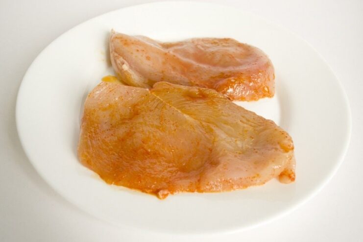 Marinating the chicken fillet with salt and red pepper before chopping. Raw chicken fillet on a cutting board being seasoned with spices.