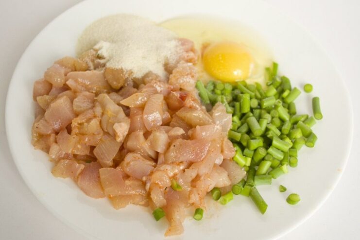 Hand-chopping chicken with egg, semolina, and garlic scapes on a board. A mixture of chopped chicken, egg, and herbs being minced with a knife on a wooden board.
