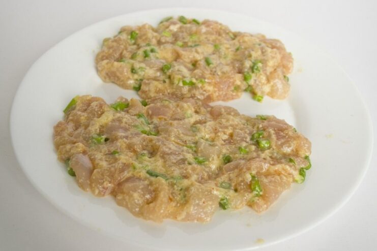 Forming the loose chicken mixture into a cutlet on a hot frying pan. Spoonfuls of raw minced chicken mixture being placed into a hot oiled frying pan.