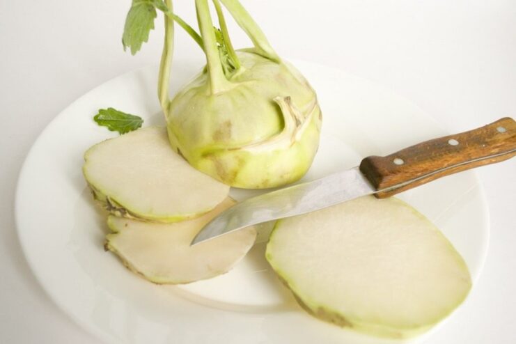 Slicing a peeled kohlrabi into thick rounds for frying. A hand holding a knife, slicing a peeled kohlrabi bulb on a cutting board.