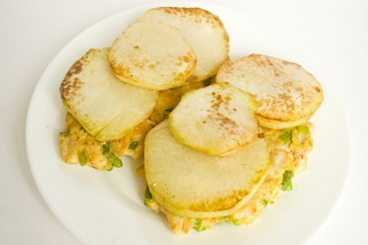Placing pan-fried kohlrabi slices on top of the cooked chicken cutlets. Golden-fried kohlrabi slices being arranged on top of chicken cutlets in a pan.