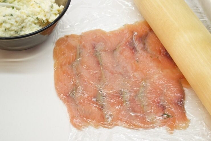 Thinly sliced cured pink salmon arranged in a rectangle on a sheet of plastic wrap.