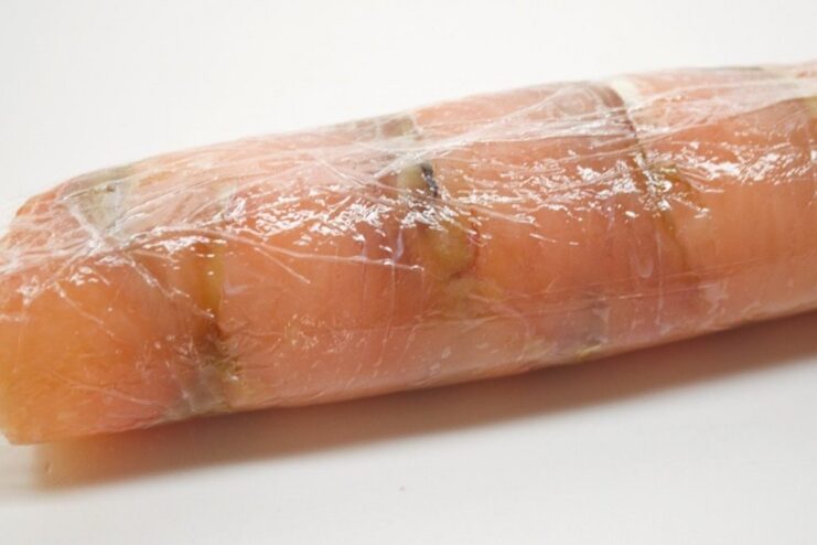 A tight roll being formed from the layer of cured salmon and cheese filling using plastic wrap.