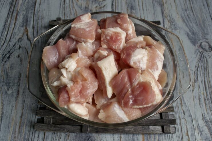 Cutting fresh pork into large, uniform chunks for the shashlik. Raw pork being cut into large cubes on a cutting board.