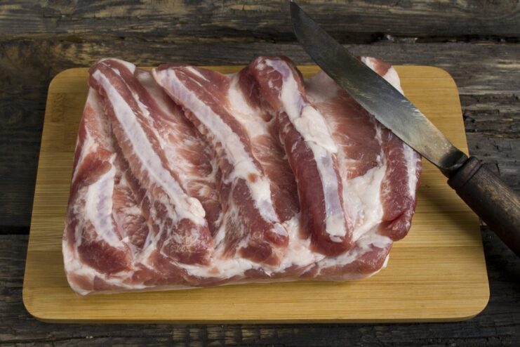 A piece of raw pork belly being cut into large sections on a cutting board.