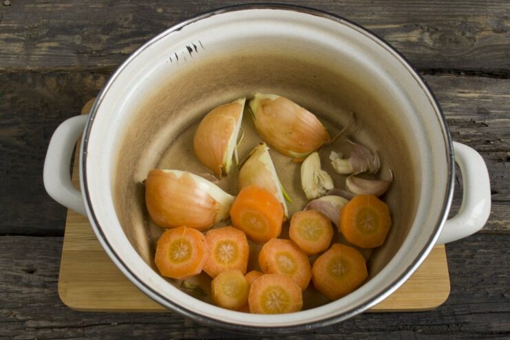 Sliced carrots, onions, and whole garlic cloves at the bottom of a large cooking pot.