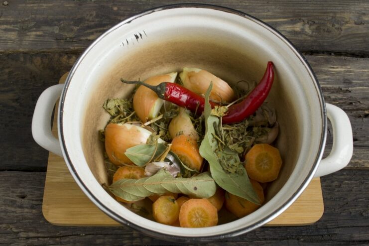 A pot with vegetables, with dried herbs, bay leaves, and a chili pepper being added.