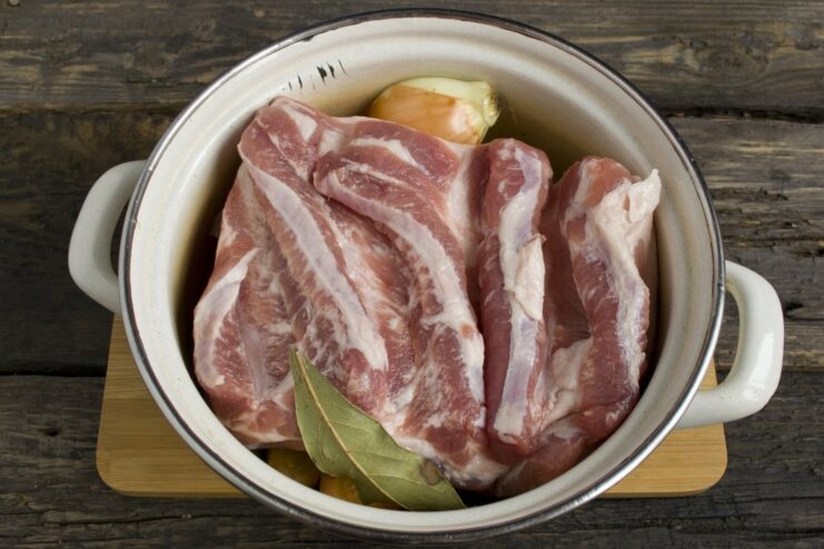 Pieces of raw pork belly placed on top of a bed of vegetables and spices in a pot.